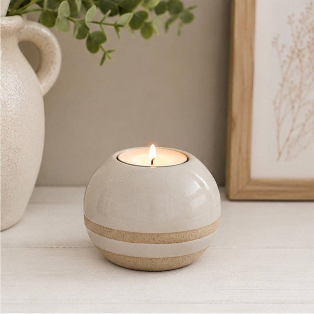 A small round ceramic candle holder with a lit tea light candle, featuring a glossy off-white top and a speckled beige band around the middle, styled on a white tabletop with a ceramic vase and framed art in the background.