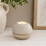 A small round ceramic candle holder with a lit tea light candle, featuring a glossy off-white top and a speckled beige band around the middle, styled on a white tabletop with a ceramic vase and framed art in the background.