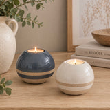 Two round ceramic tea light holders on a wooden table, one in dark blue-gray and one in warm off-white, both with a sandy beige band and lit with tea light candles. Decorative props include a ceramic vase with eucalyptus, stacked books, and a framed print in the background.