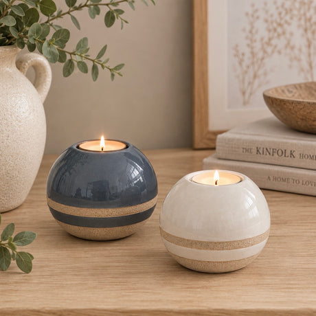 Two round ceramic tea light holders on a wooden table, one in dark blue-gray and one in warm off-white, both with a sandy beige band and lit with tea light candles. Decorative props include a ceramic vase with eucalyptus, stacked books, and a framed print in the background.