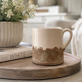 A ceramic jug with a textured beige upper half and a rustic brown glazed lower half, resting on a round wooden tray next to a book and a potted plant.