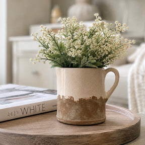 A beige ceramic jug with a rustic brown glazed base, filled with white baby's breath flowers, displayed on a round wooden tray next to a book in a softly blurred living room setting.