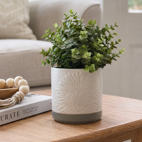 A potted artificial green plant in a white ceramic pot with an embossed sunflower design and a grey base, placed on a wooden table in a softly lit living room setting.