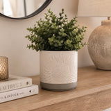 An artificial green plant in a white cylindrical planter with an embossed sunflower design and a grey base, displayed on a wooden console table next to a stack of books, a candle, and a lamp.