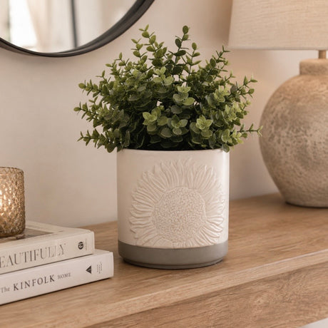 An artificial green plant in a white cylindrical planter with an embossed sunflower design and a grey base, displayed on a wooden console table next to a stack of books, a candle, and a lamp.