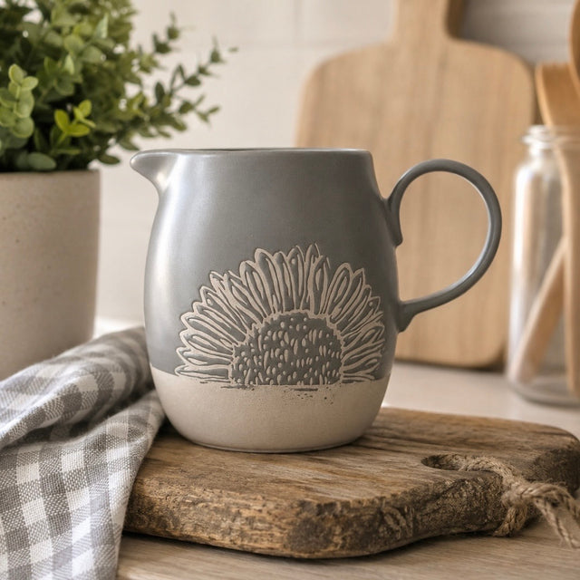 A grey ceramic pitcher with a sunflower design, resting on a rustic wooden cutting board next to a checkered cloth napkin in a kitchen setting.