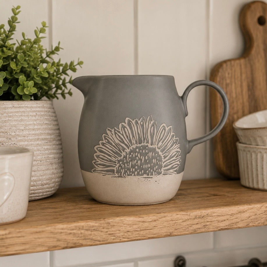 A grey ceramic pitcher with a beige sunflower design, sitting on a wooden shelf in a kitchen setting.