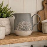 A grey ceramic pitcher with a beige sunflower design, sitting on a wooden shelf in a kitchen setting.