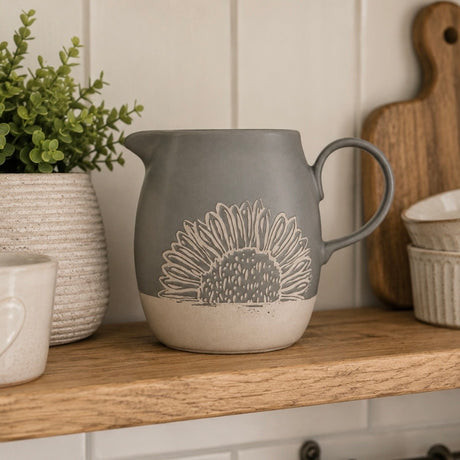 A grey ceramic pitcher with a beige sunflower design, sitting on a wooden shelf in a kitchen setting.