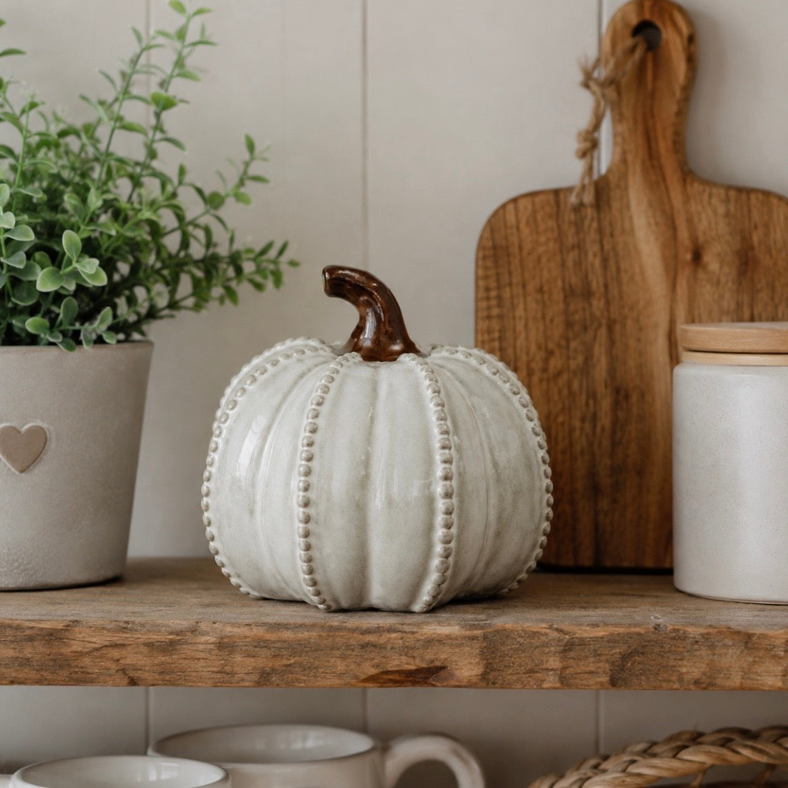 A decorative ceramic pumpkin in a warm off-white glaze with a dark brown stem, styled on a rustic wooden shelf alongside a potted green plant, a wooden cutting board, and ceramic canisters.