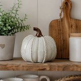 A decorative ceramic pumpkin in a warm off-white glaze with a dark brown stem, styled on a rustic wooden shelf alongside a potted green plant, a wooden cutting board, and ceramic canisters.