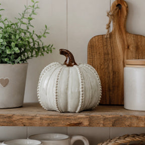A decorative ceramic pumpkin in a warm off-white glaze with a dark brown stem, styled on a rustic wooden shelf alongside a potted green plant, a wooden cutting board, and ceramic canisters.