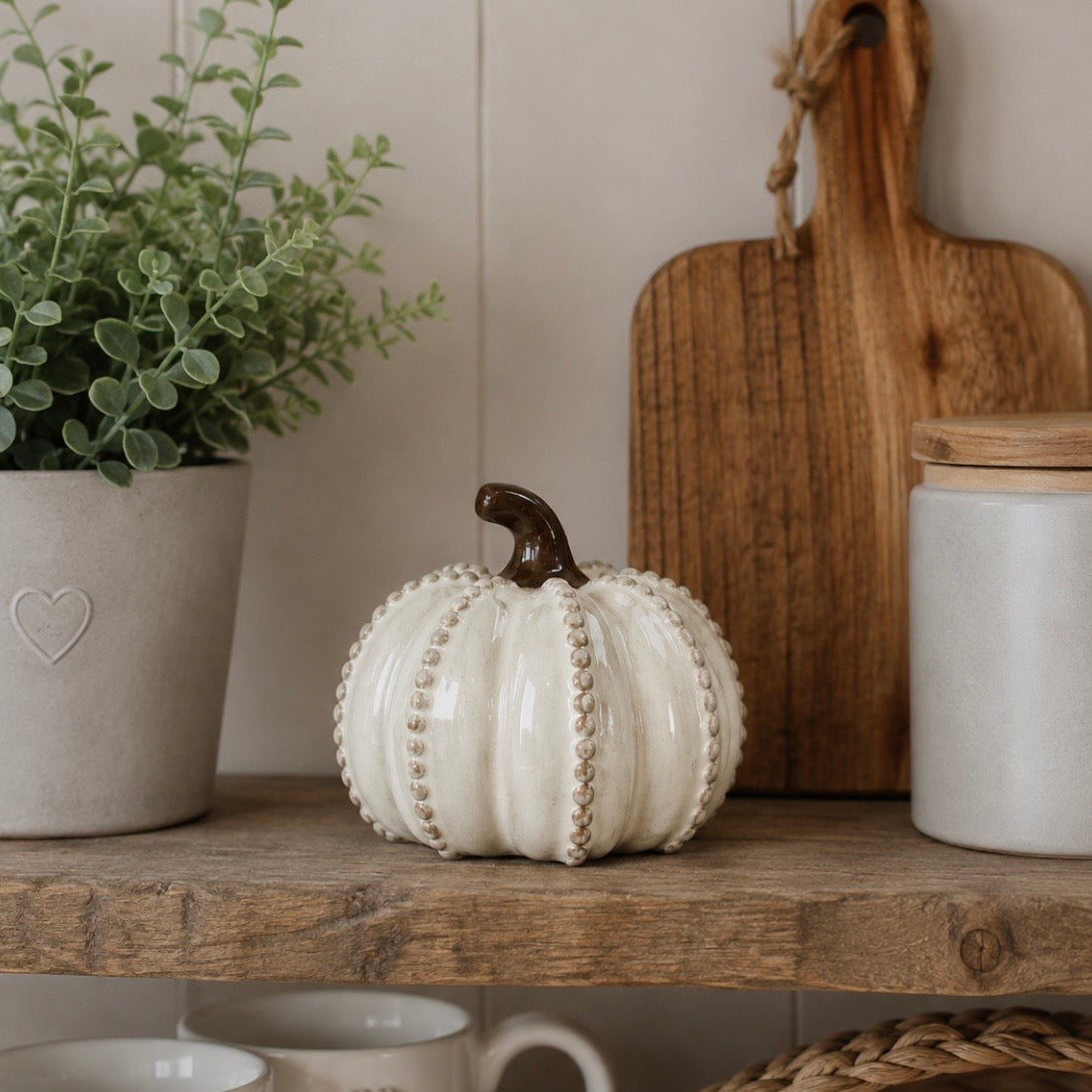 A decorative ceramic pumpkin in off-white with a dark brown stem sits on a rustic wooden shelf, styled with a potted green plant, a wooden cutting board, and a white jar in a home interior setting.