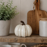 A decorative ceramic pumpkin in off-white with a dark brown stem sits on a rustic wooden shelf, styled with a potted green plant, a wooden cutting board, and a white jar in a home interior setting.
