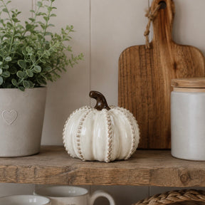 A decorative ceramic pumpkin in off-white with a dark brown stem sits on a rustic wooden shelf, styled with a potted green plant, a wooden cutting board, and a white jar in a home interior setting.