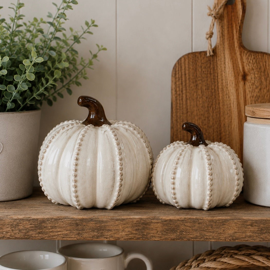 Two decorative ceramic pumpkins in off-white with beaded trim and dark brown stems, displayed on a wooden shelf in a styled home setting with a potted plant, cutting board, and mugs.