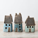 Three small ceramic house figurines in blue and turquoise glazes with dark brown roofs, displayed on a wooden shelf surface
