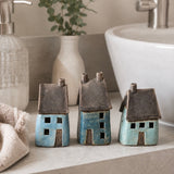 Three small ceramic house figurines in blue and turquoise glazes with dark brown roofs, displayed on a marble surface next to a white ceramic sink in a bathroom scene