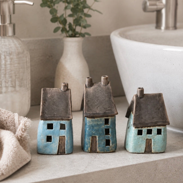 Three small ceramic house figurines in blue and turquoise glazes with dark brown roofs, displayed on a marble surface next to a white ceramic sink in a bathroom scene
