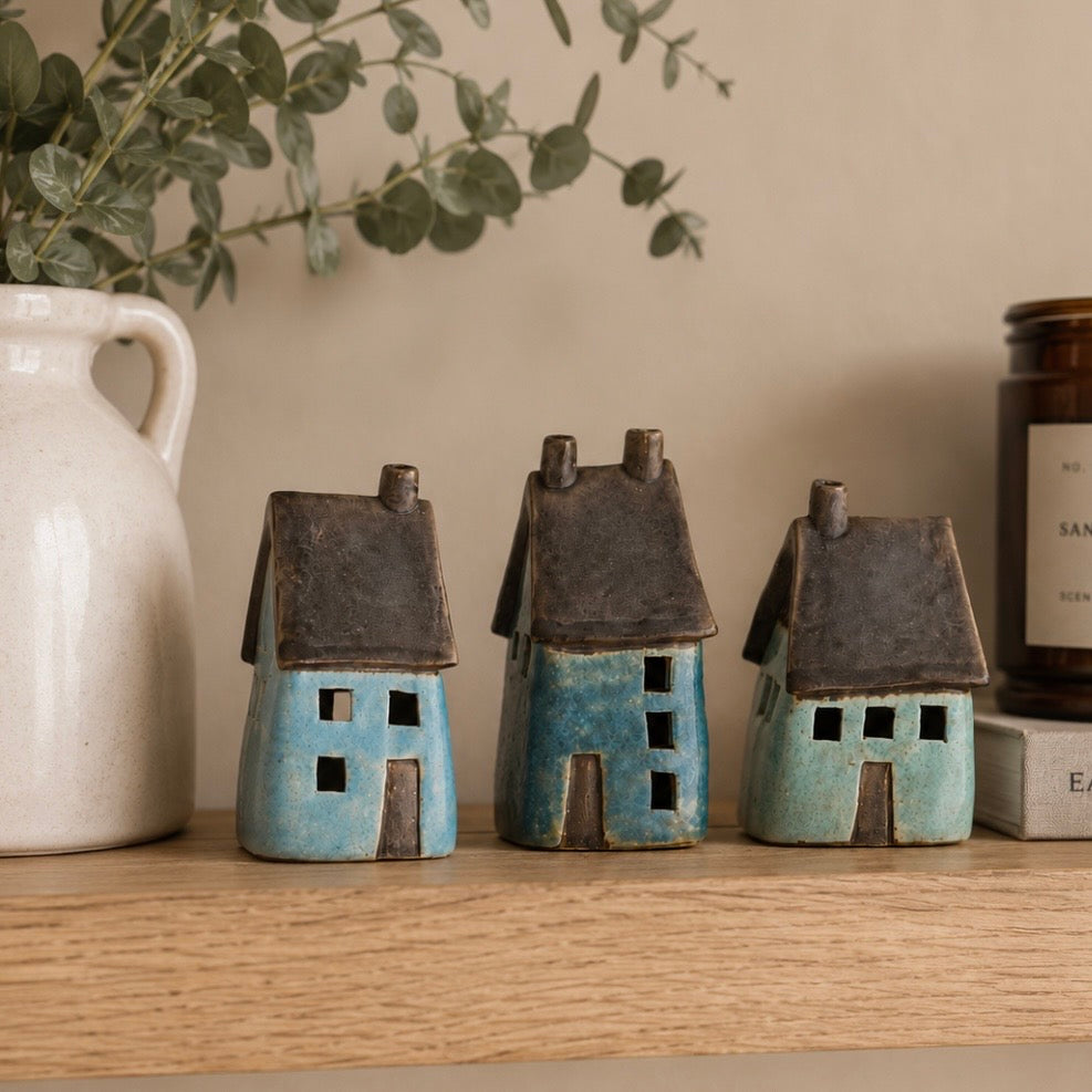 Three small ceramic house figurines in blue and turquoise glazes with dark brown roofs, displayed on a wooden shelf next to a white ceramic pitcher and a candle.