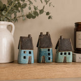 Three small ceramic house figurines in blue and turquoise glazes with dark brown roofs, displayed on a wooden shelf next to a white ceramic pitcher and a candle.