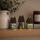Three small ceramic LED house figurines in blue and turquoise glazes with dark brown roofs, displayed on a wooden shelf next to a white ceramic pitcher and a candle.