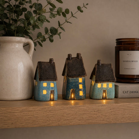 Three small ceramic LED house figurines in blue and turquoise glazes with dark brown roofs, displayed on a wooden shelf next to a white ceramic pitcher and a candle.