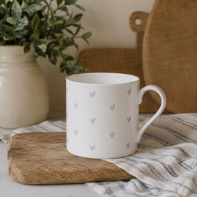 A white fine bone china mug with a pattern of small grey hearts, resting on a wooden cutting board over a striped cloth. A potted green plant and a wooden cutting board are blurred in the background.