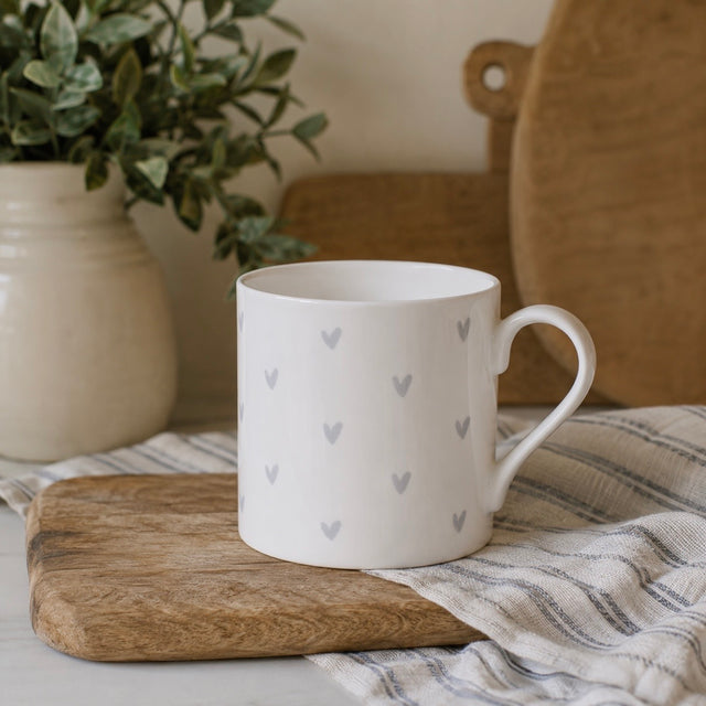 A white fine bone china mug with a pattern of small grey hearts, resting on a wooden cutting board over a striped cloth. A potted green plant and a wooden cutting board are blurred in the background.