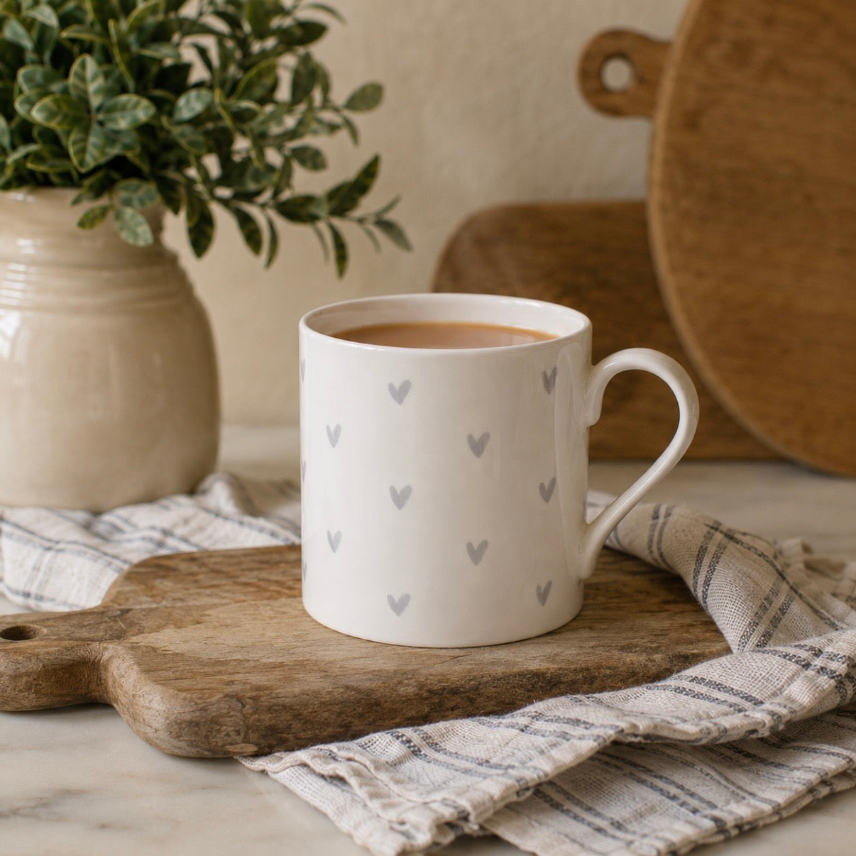 A white fine bone china mug with a pattern of small grey hearts, resting on a wooden cutting board over a striped cloth. A potted green plant and a wooden cutting board are blurred in the background.