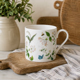 A white fine bone china mug decorated with delicate botanical illustrations of flowers, leaves, a bee, and a butterfly, styled on a wooden cutting board with a striped cloth and a potted plant in the background.
