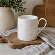 A white fine bone china mug with a handle, decorated with a pattern of small pink hearts, resting on a wooden cutting board with a striped cloth underneath.