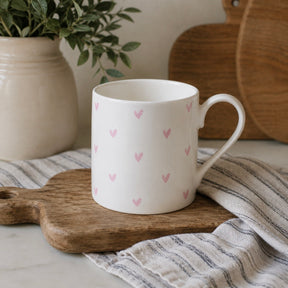 A white fine bone china mug with a handle, decorated with a pattern of small pink hearts, resting on a wooden cutting board with a striped cloth underneath.