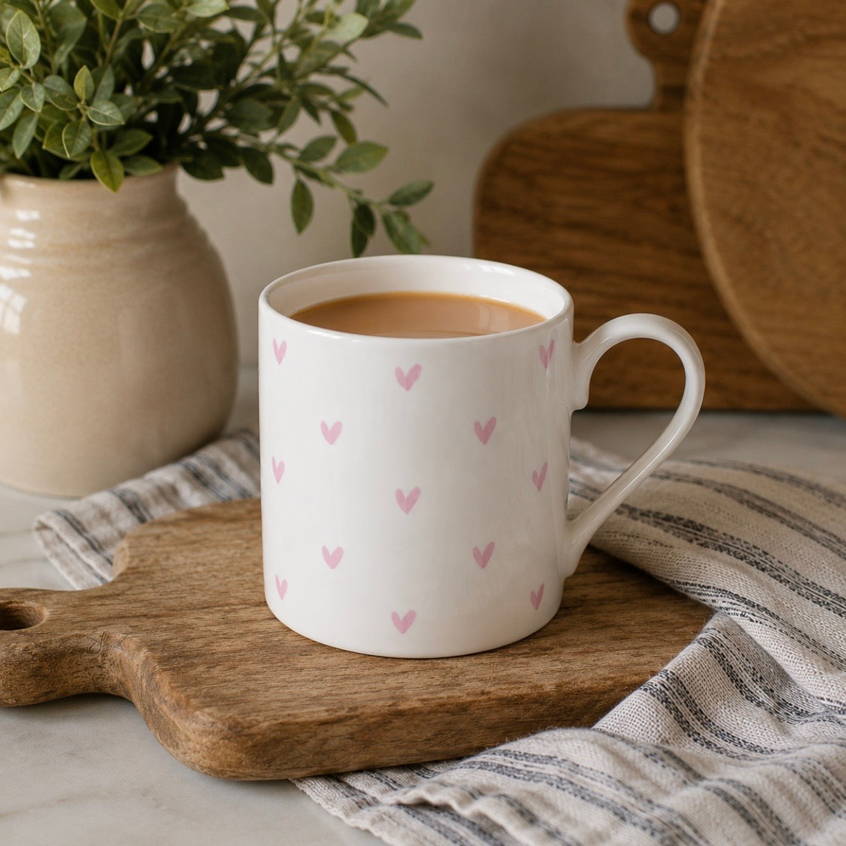 A white fine bone china mug with a handle, decorated with a pattern of small pink hearts, resting on a wooden cutting board with a striped cloth underneath.