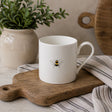 A white fine bone china mug with a handle, decorated with a single bee, resting on a wooden cutting board with a striped cloth underneath.