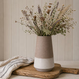 A two-tone ceramic vase with a textured matte finish, featuring a warm mocha brown upper half and a cream lower half, filled with dried flowers and placed on a rustic wooden board with a striped cloth against a beige paneled wall.