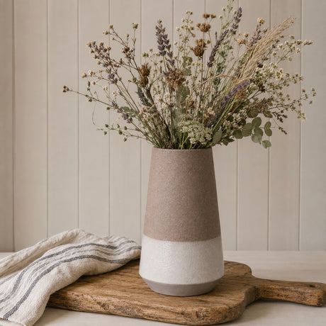 A two-tone ceramic vase with a textured matte finish, featuring a warm mocha brown upper half and a cream lower half, filled with dried flowers and placed on a rustic wooden board with a striped cloth against a beige paneled wall.