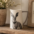 A white ceramic pitcher with a grey handle, featuring a detailed black and white illustration of a hare on the front. The pitcher is styled on a rustic wooden shelf next to a potted daisy and a wooden picture frame.