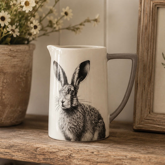 A white ceramic pitcher with a grey handle, featuring a detailed black and white illustration of a hare on the front. The pitcher is styled on a rustic wooden shelf next to a potted daisy and a wooden picture frame.