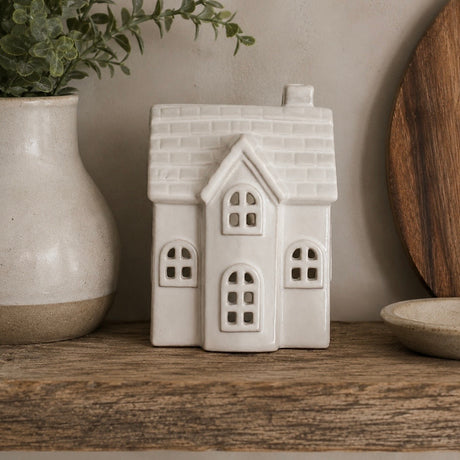A small white ceramic house-shaped decorative object with arched windows and a textured roof, displayed on a wooden surface beside a vase with greenery and a round wooden board.
