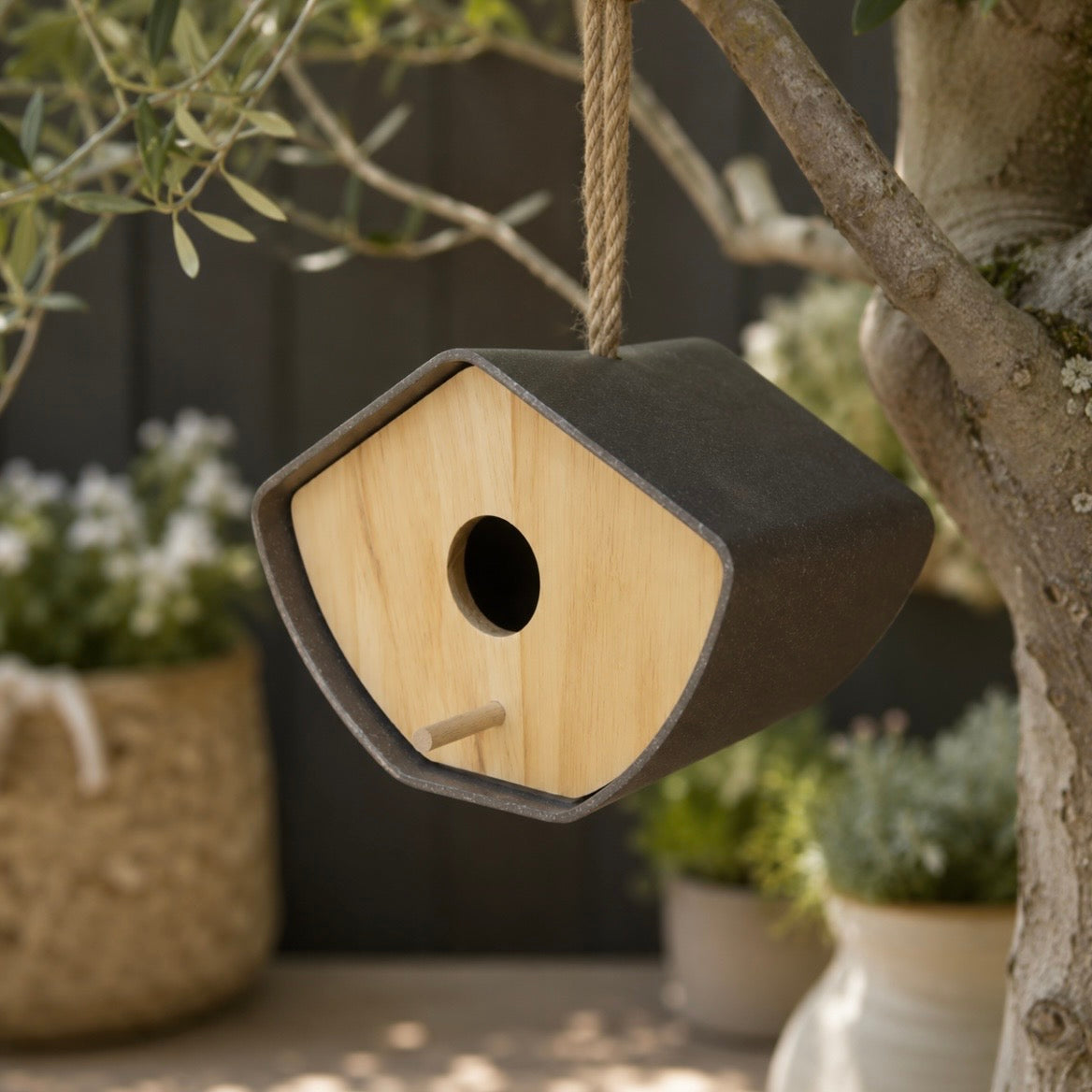 A small birdhouse hanging from a rope in an outdoor garden setting, with a dark charcoal exterior and a light natural wood front panel.