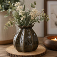 A dark green glossy ceramic vase with a poppy bud shape, filled with eucalyptus and small white flowers, displayed on a round wooden slice coaster with a warm candle in the background.
