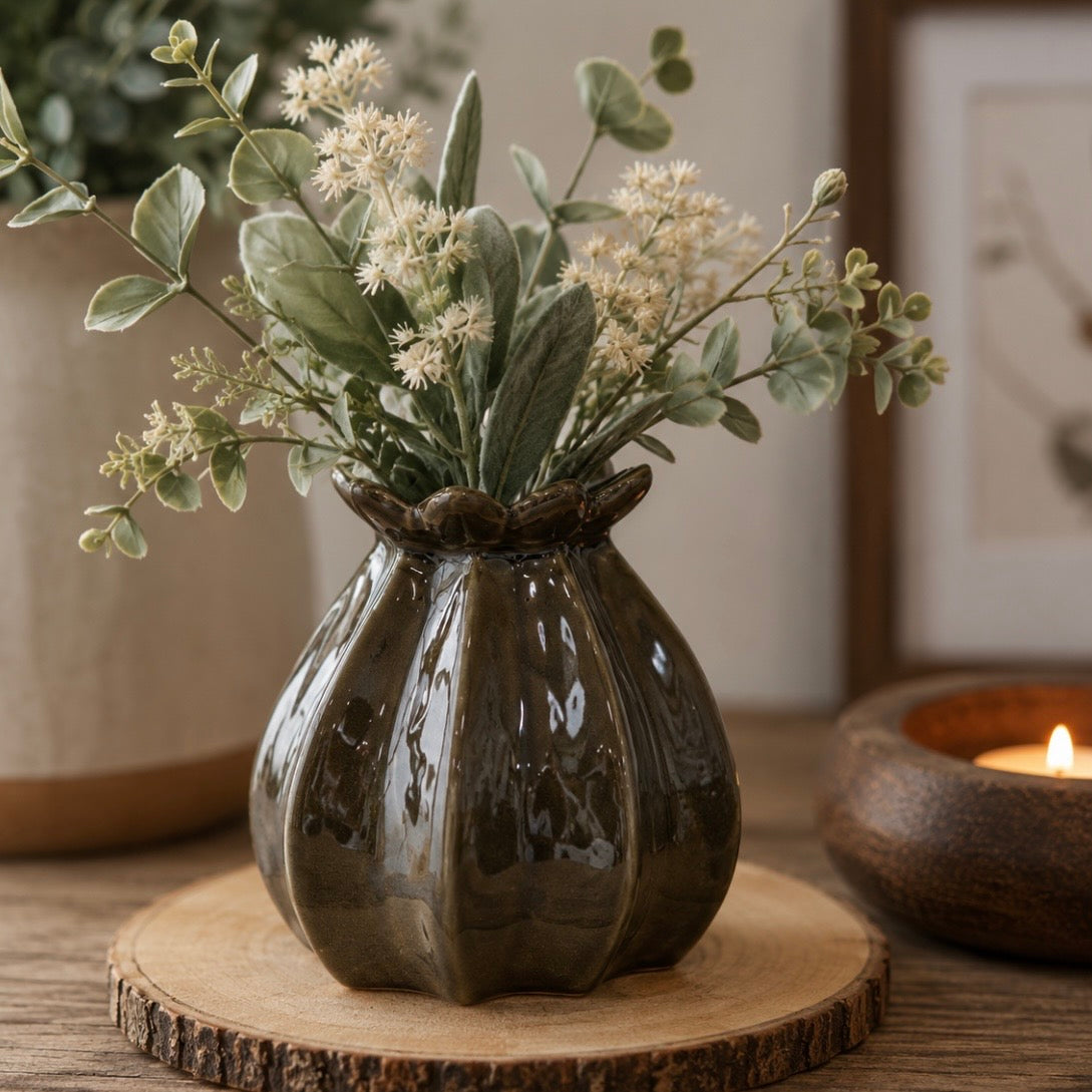 A dark green glossy ceramic vase with a poppy bud shape, filled with eucalyptus and small white flowers, displayed on a round wooden slice coaster with a warm candle in the background.
