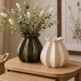Two decorative ceramic poppy bud-shaped vases on a wooden table, one dark green and one cream, with dried grasses and eucalyptus stems in the larger vase, in a softly styled interior setting.