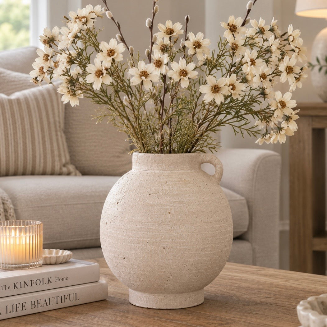 A round, textured off-white terracotta vase with a side loop handle, filled with white daisy-like dried flowers and green stems, placed on a wooden table in a cozy living room setting.