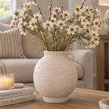 A round, textured off-white terracotta vase with a side loop handle, filled with white daisy-like dried flowers and green stems, placed on a wooden table in a cozy living room setting.