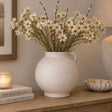 A textured cream terracotta vase filled with white daisy-like dried flowers and brown stems, displayed on a wooden console table in a styled room setting with a candle, books, and a lamp.