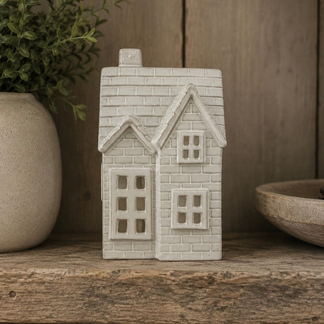 An LED House shaped tealight in an off-white glaze, displayed on a rustic wooden surface with a potted plant and bowl in the background.