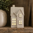 A House shaped ceramic LED tealight with a warm yellow glow from within, displayed on a rustic wooden shelf next to a potted plant.