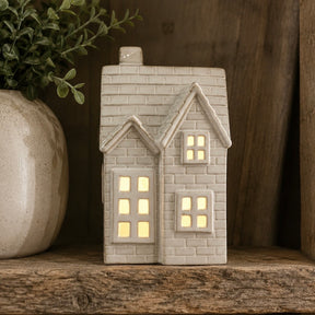 A House shaped ceramic LED tealight with a warm yellow glow from within, displayed on a rustic wooden shelf next to a potted plant.